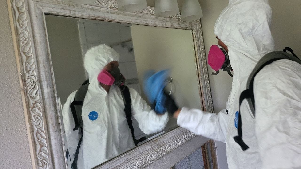 Mold remediation specialist in PPE wiping down bathroom mirror with blue cloth during mold cleanup
