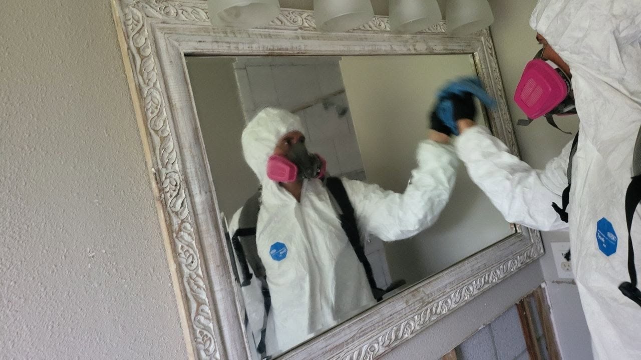 Two mold remediation technicians in full PPE suits cleaning bathroom mirror during mold removal