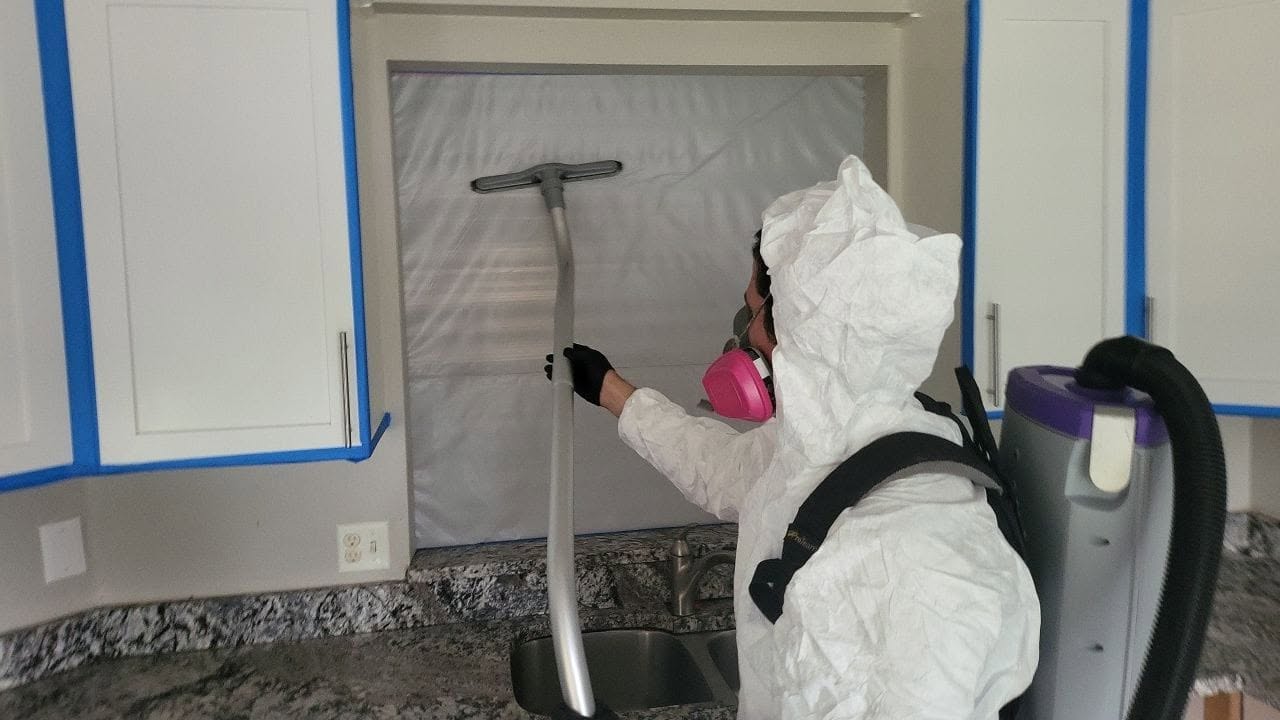Mold remediation specialist in PPE with HEPA vacuum working at plastic containment barrier in kitchen