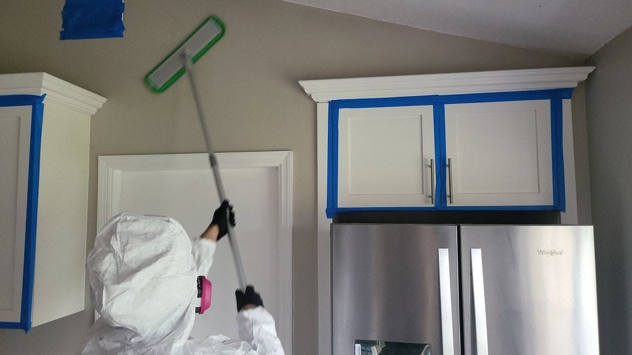 Mold remediation technician in full PPE mopping ceiling above kitchen cabinets sealed with blue tape containment