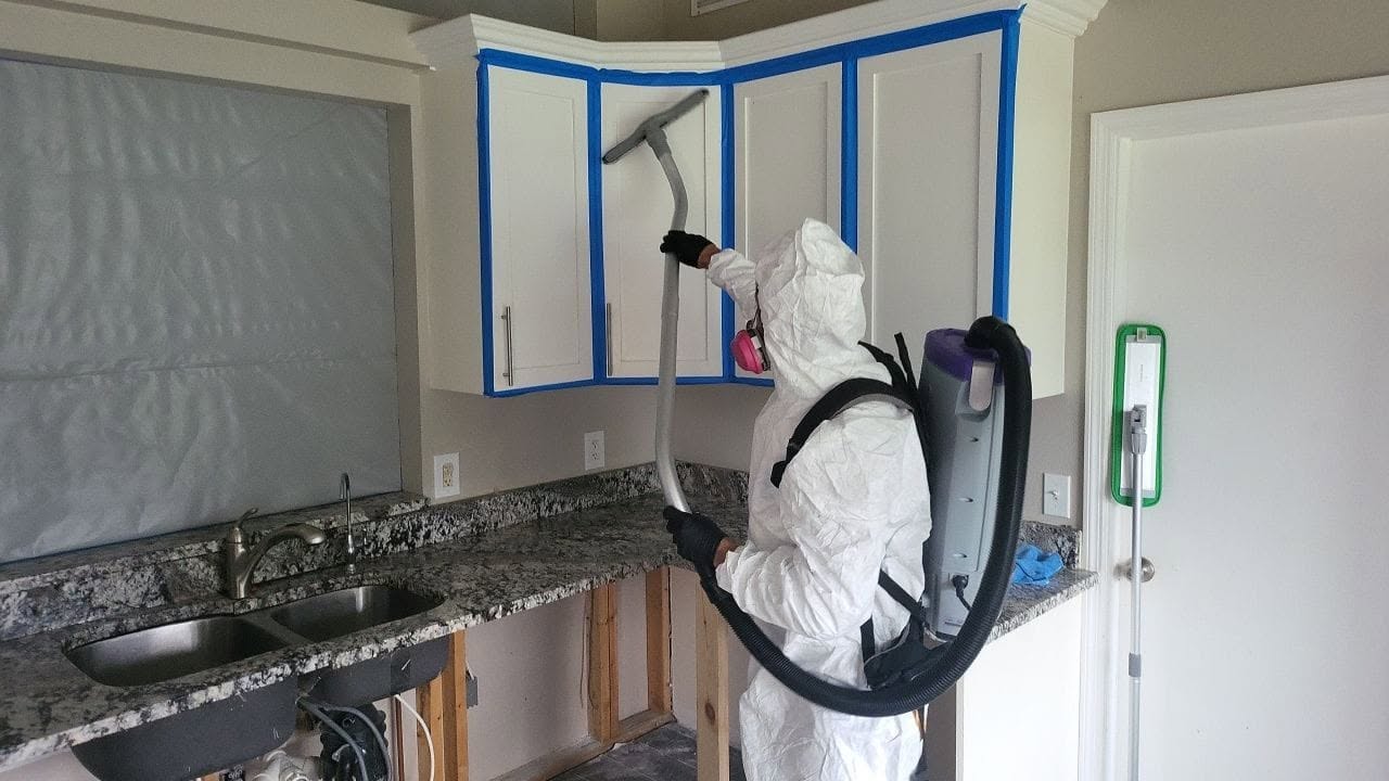 Mold remediation technician in Tyvek suit using backpack HEPA vacuum on kitchen cabinets with blue tape containment