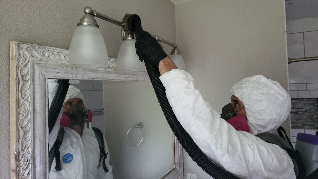 Mold remediation technician in full PPE suit vacuuming bathroom light fixture during mold cleanup