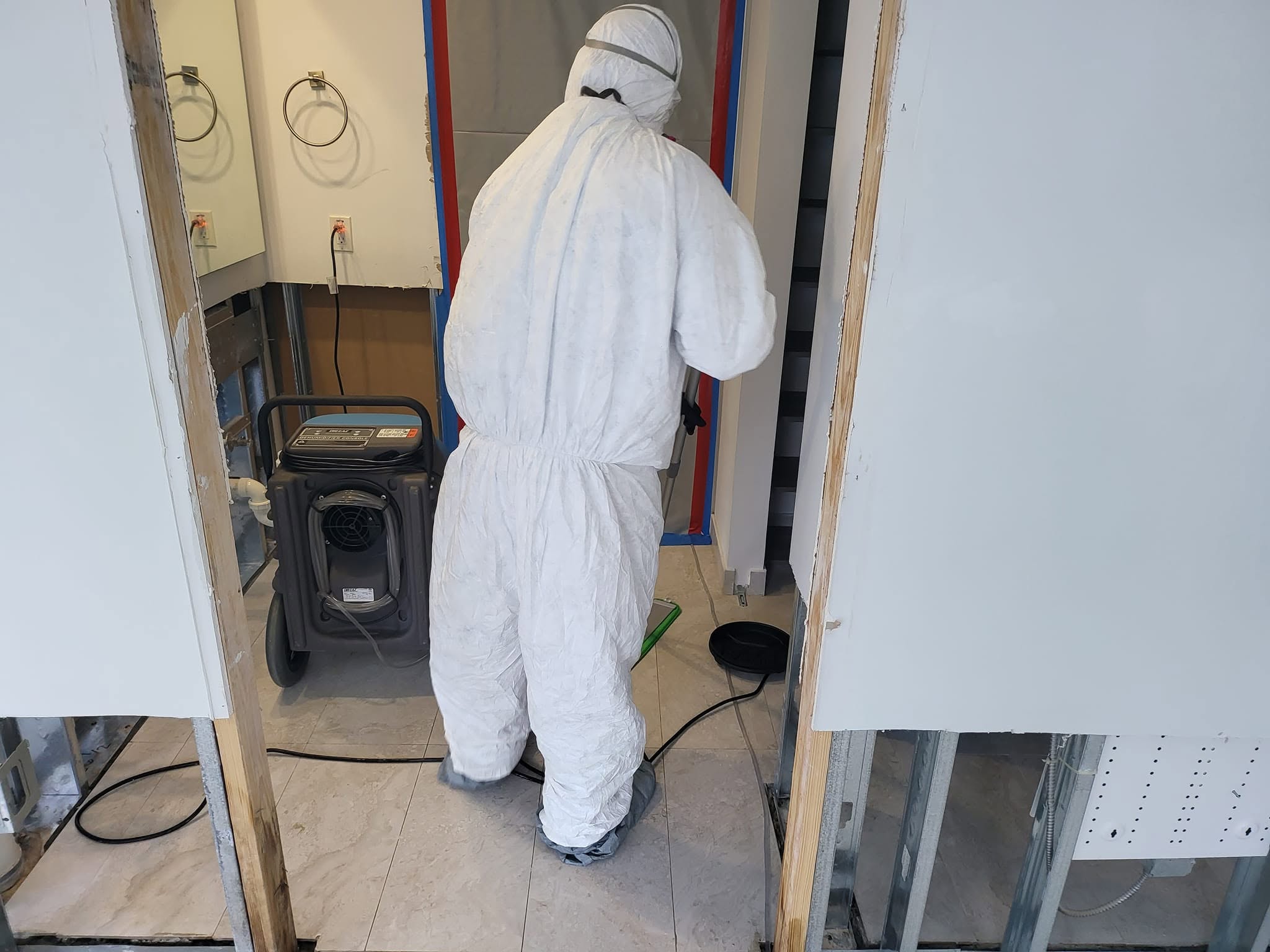 Mold remediation technician in Tyvek suit working in stripped bathroom doorway with dehumidifier running