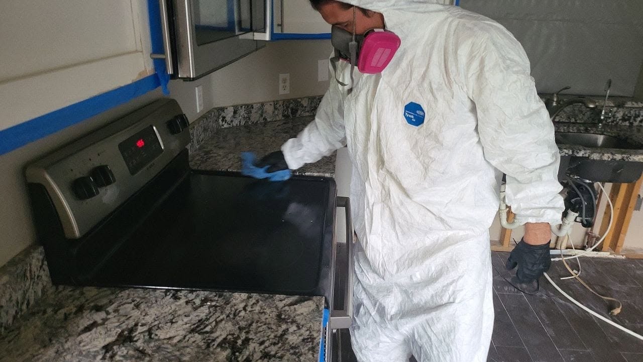 Mold remediation technician in Tyvek suit wiping down kitchen countertop and stove during mold cleanup