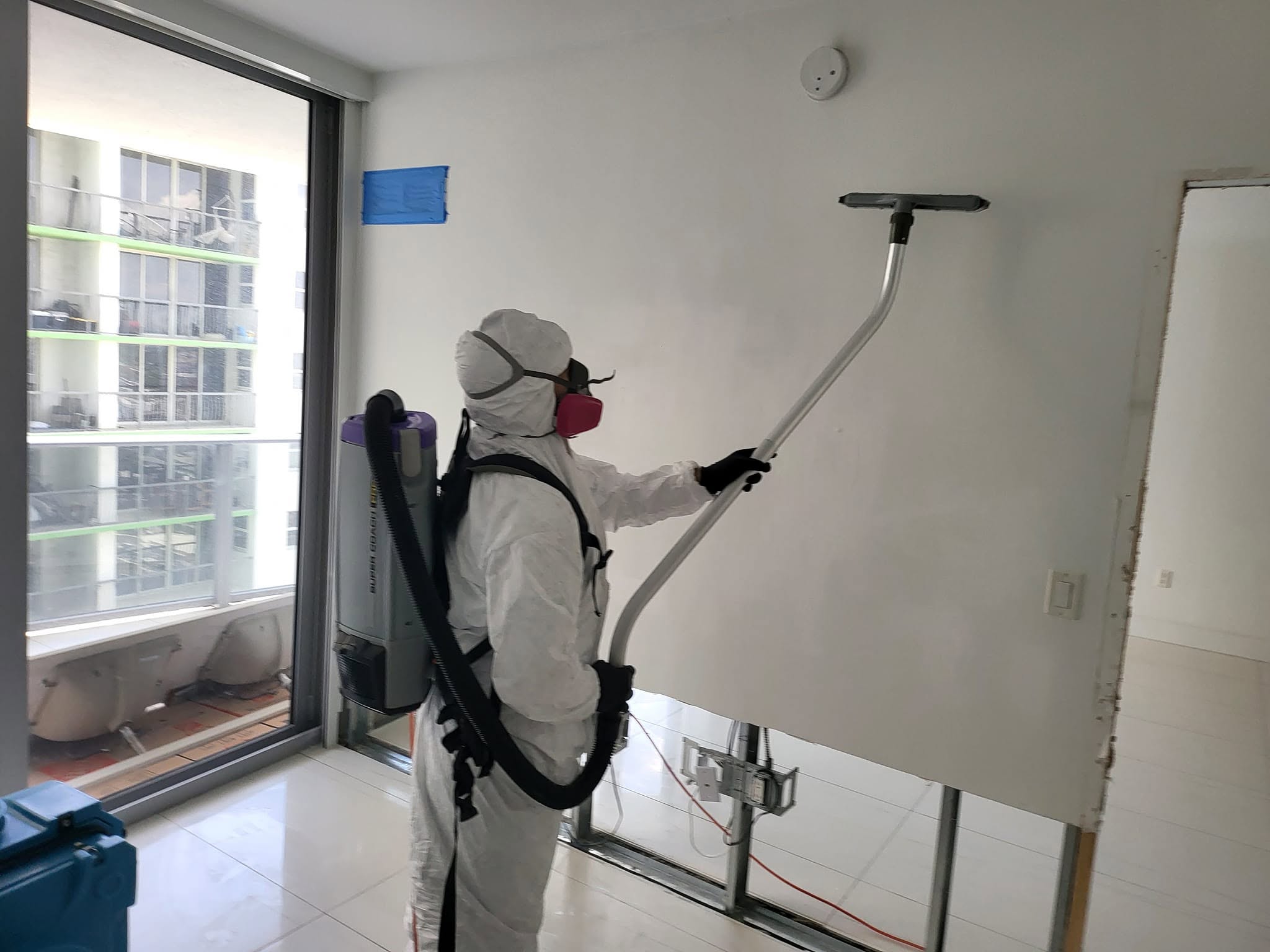 Mold remediation technician in full Tyvek suit with backpack HEPA vacuum wiping condo wall during mold cleanup
